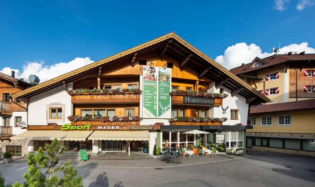 Alpines Hotel Hubertushof mit Holzfassade, Blumenschmuck auf den Balkonen und Sportgeschäft Sport Mader im Erdgeschoss.