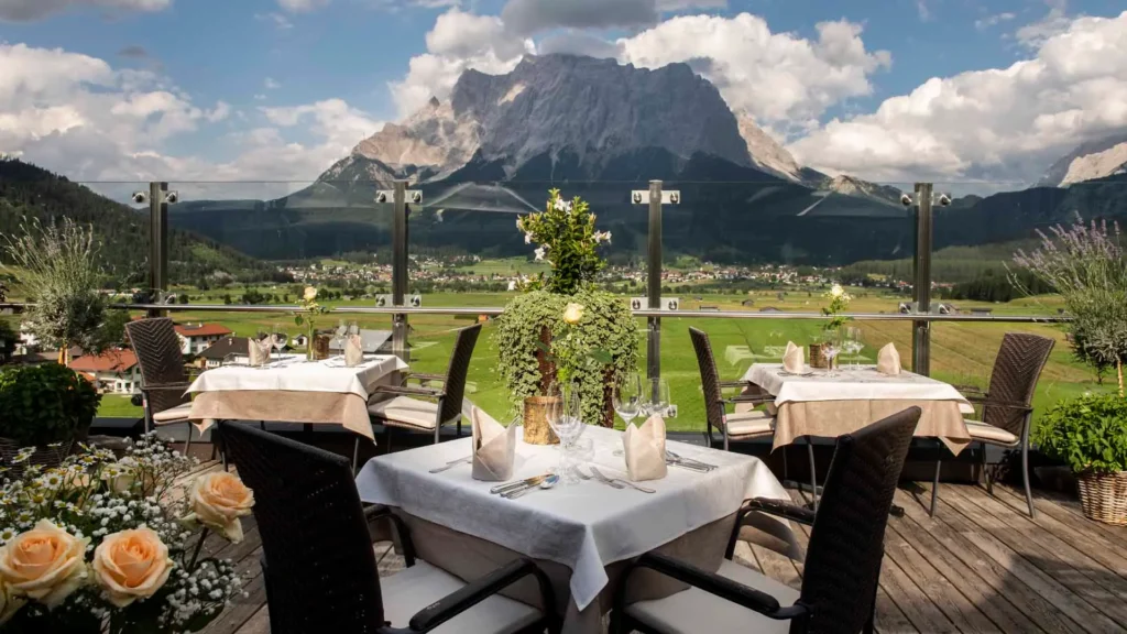Eingedeckte Tische auf der Holzterrasse des Hotel Hubertushof mit Blumenschmuck und freiem Blick auf die Zugspitze.