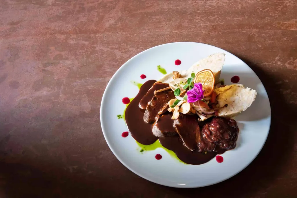 Zartes Bratenfleisch mit dunkler Soße, Knödeln und essbarer Blütendeko auf weißem Teller im Hotel Hubertushof.