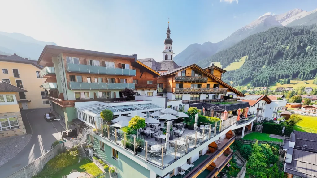 Außenansicht des Hotel Hubertushof mit sonniger Dachterrasse, Wintergarten und Kirchturm vor Bergkulisse.