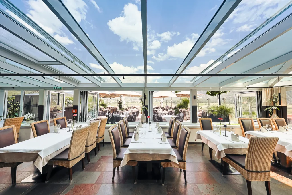 Lichtdurchfluteter Wintergarten im Hotel Hubertushof mit eingedeckten Tischen und Blick auf die Terrasse.