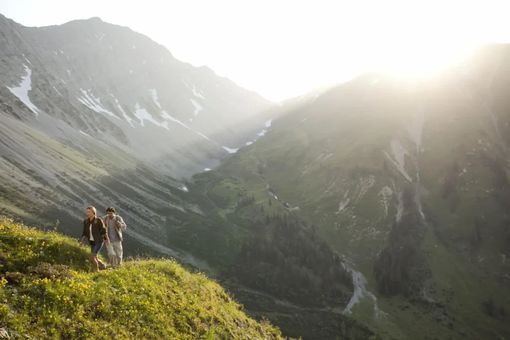 Zwei Wanderer steigen im Sonnenlicht über eine blühende Bergwiese mit Panorama für Gäste des Hotel Hubertushof.